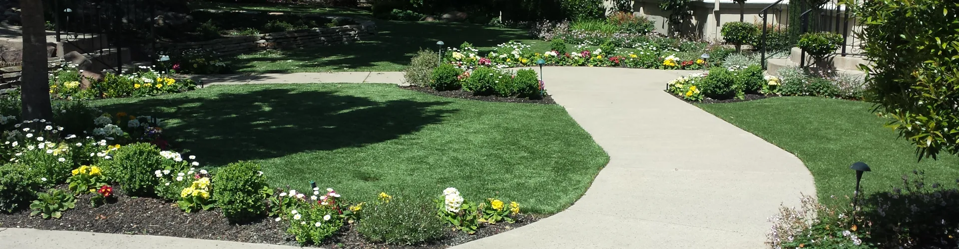 Artificial turf front yard with a curved concrete walkway, colorful flower beds, topiary shrubs, and a Spanish-style stucco home with red tile roof