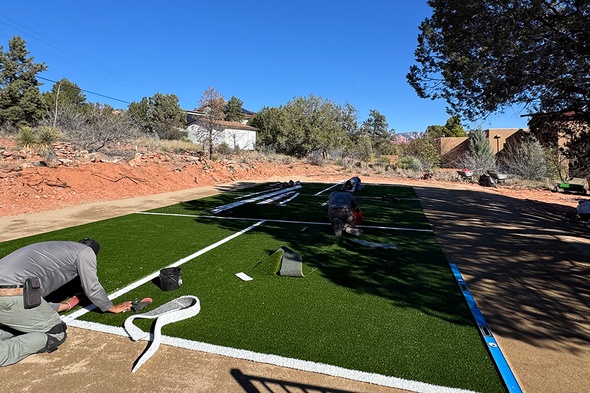 Installers laying and seaming synthetic grass installation for a pickleball court with white boundary lines in a Sedona, Arizona backyard bordered by natural red rock formations and desert vegetation.
